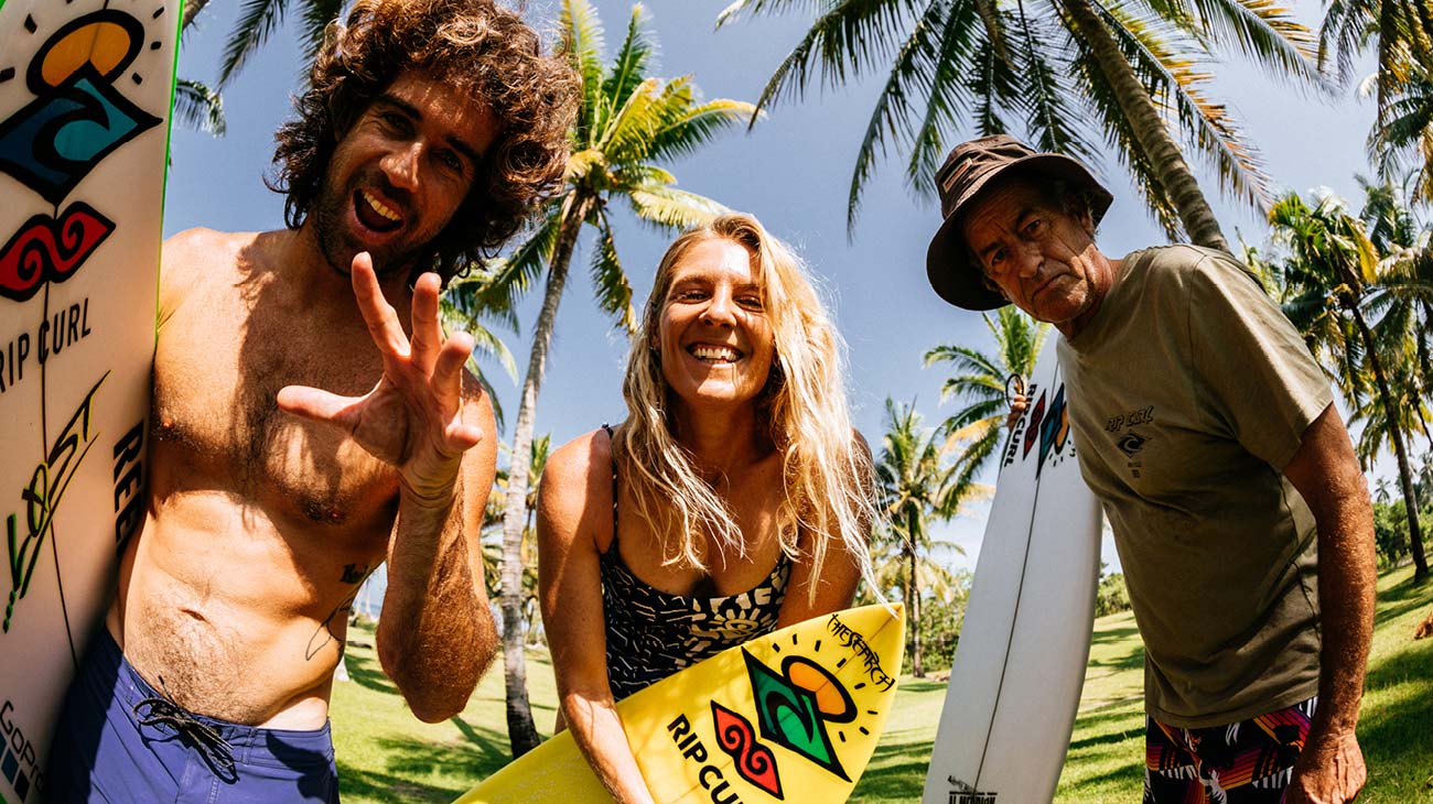 Three people with surfboards posing in a tropical setting with palm trees.