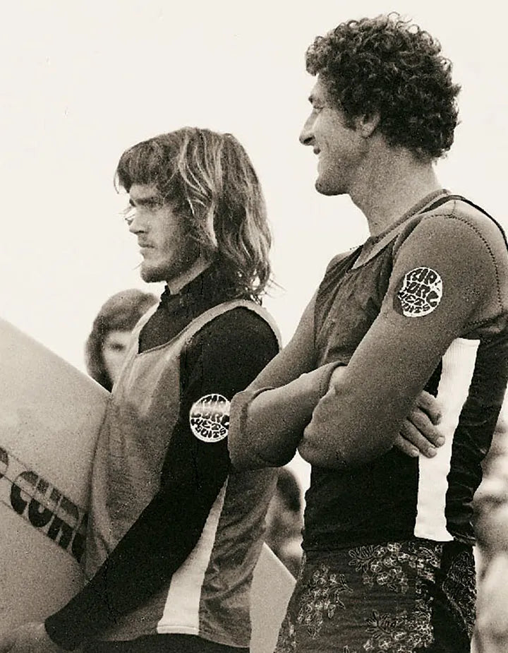Two men standing next to each other with surfboards, wearing sleeveless shirts with logos.
