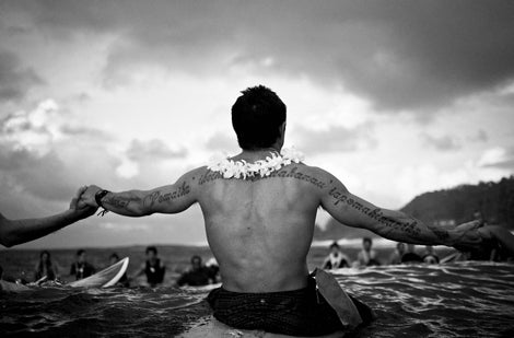 Man with tattoos and a lei on a beach with people and boats in the background
