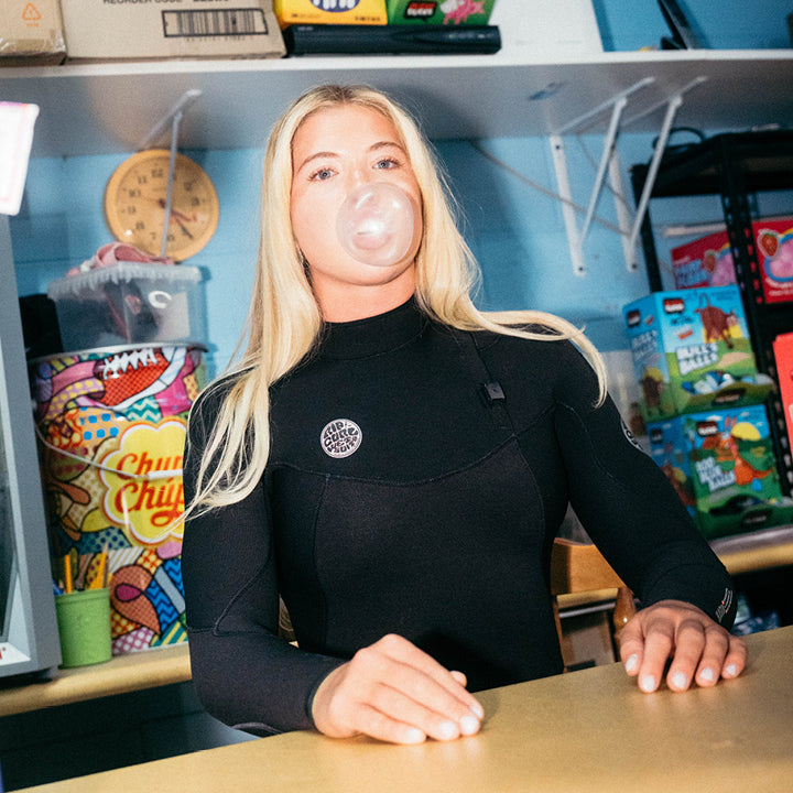 A blonde woman wearing a black long-sleeve wetsuit sits behind a counter in a shop, blowing a bubble gum bubble. Shelves in the background display various items including a Chupa Chups container and boxed toys.