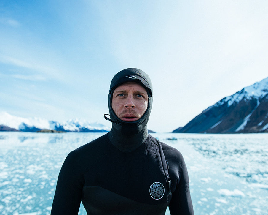 Person in a black wetsuit standing in front of a snowy landscape with mountains.