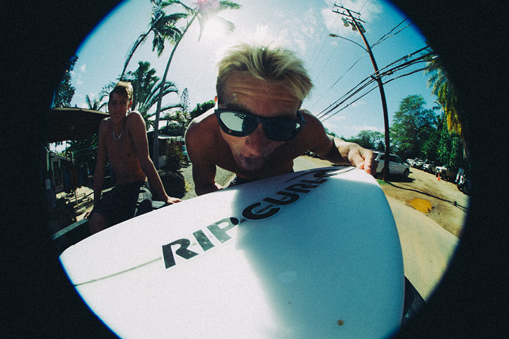 Person holding a surfboard with 'Rip Curl' branding, surrounded by palm trees and a clear sky.