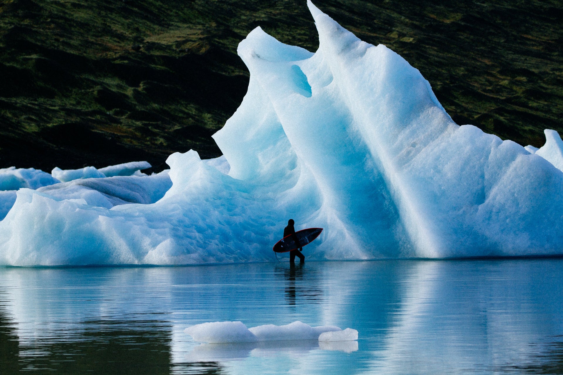 Person with a surfboard near a large iceberg in a body of water