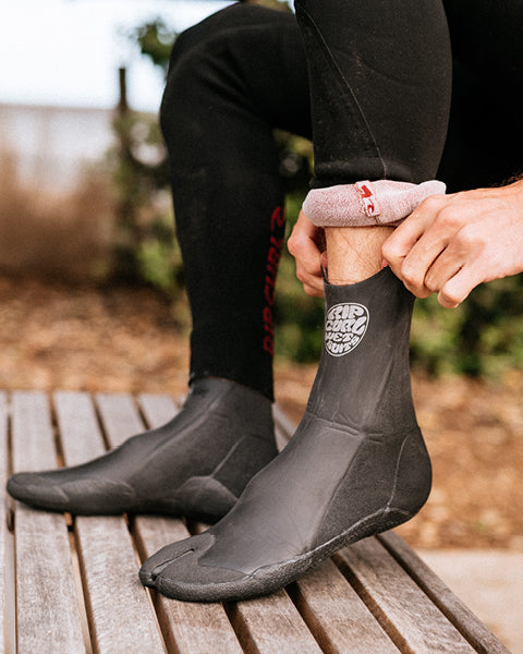 Person wearing black wetsuit boots with a visible brand logo on a wooden surface.