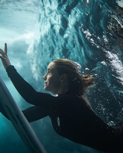 Person in a wetsuit underwater with a surfboard, surrounded by ocean waves.